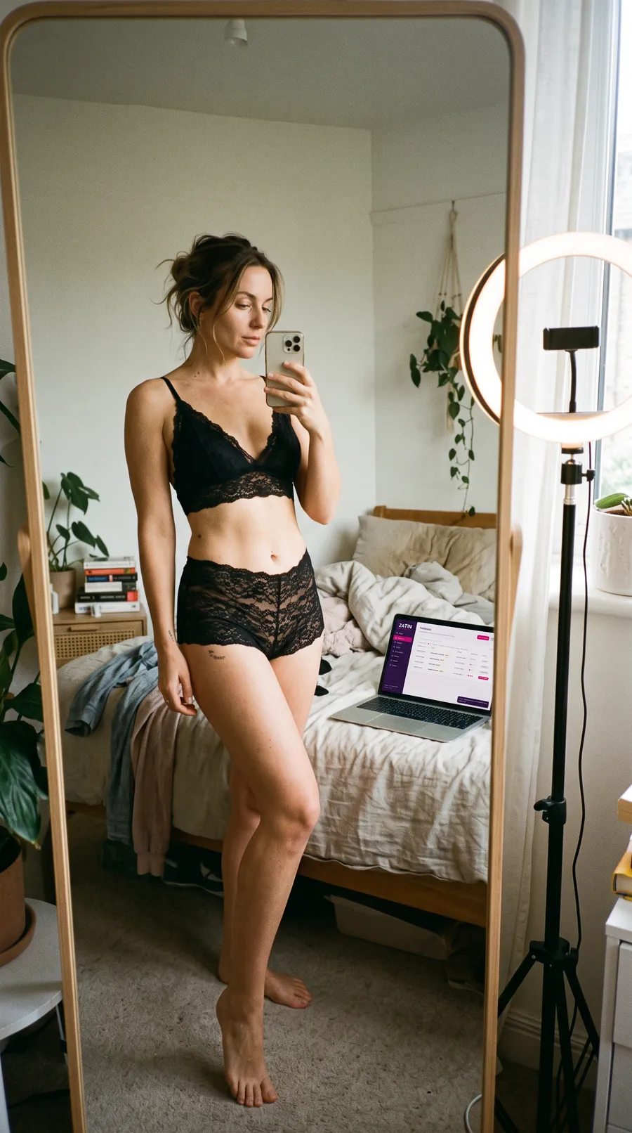A creator in black lingerie takes a mirror selfie with her phone, a ring light and a laptop with the Zatin catalogue behind her.