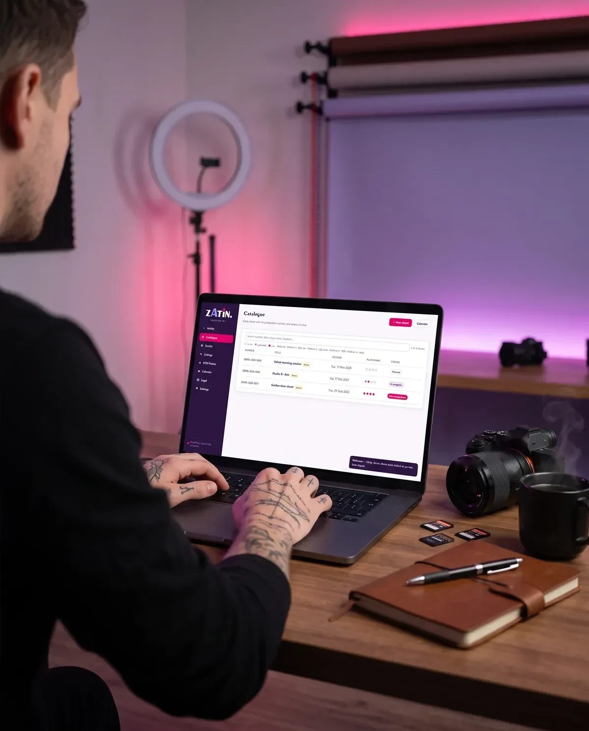 A creator at a desk, seen over the shoulder, working in the Zatin catalogue, camera and notebook beside the laptop.